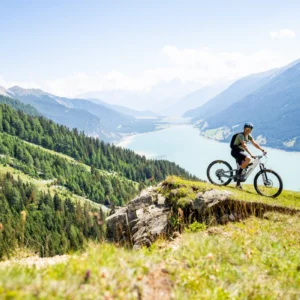 A cyclist enjoys an e-bike tour across a grassy hillside overlooking a scenic lake and forested mountains.
