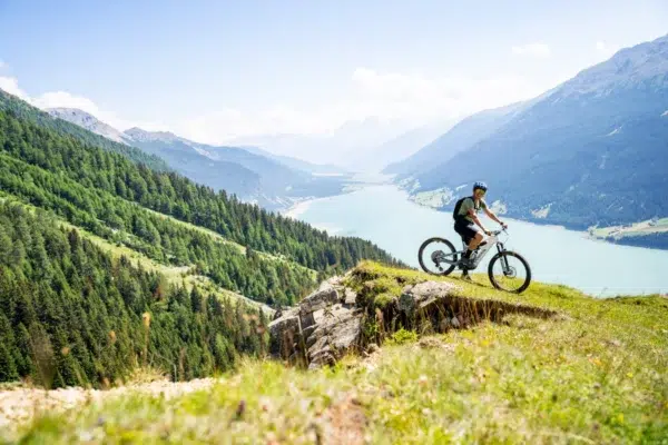 Ein Radfahrer unternimmt eine E-Bike-Tour auf einem grasbewachsenen Hügel über einem malerischen See und bewaldeten Bergen.