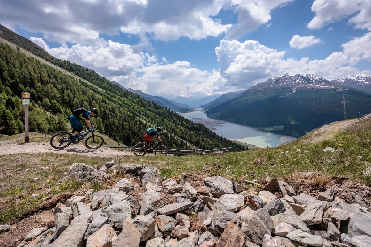 Zwei Radfahrer fahren die Genussrunde Reschensee mit Blick auf einen See und ferne Gipfel.