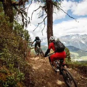 Two cyclists ride trail afternoon bikes along a dirt track through a forested mountain landscape.