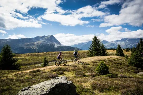 Zwei Personen fahren mit dem Mountainbike die Plamort-Tour mit Bergen und Bäumen im Hintergrund.