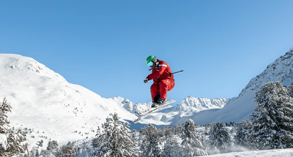 Skifahrer in rotem Anzug springt an einem klaren Tag in der Luft über eine verschneite Berglandschaft.