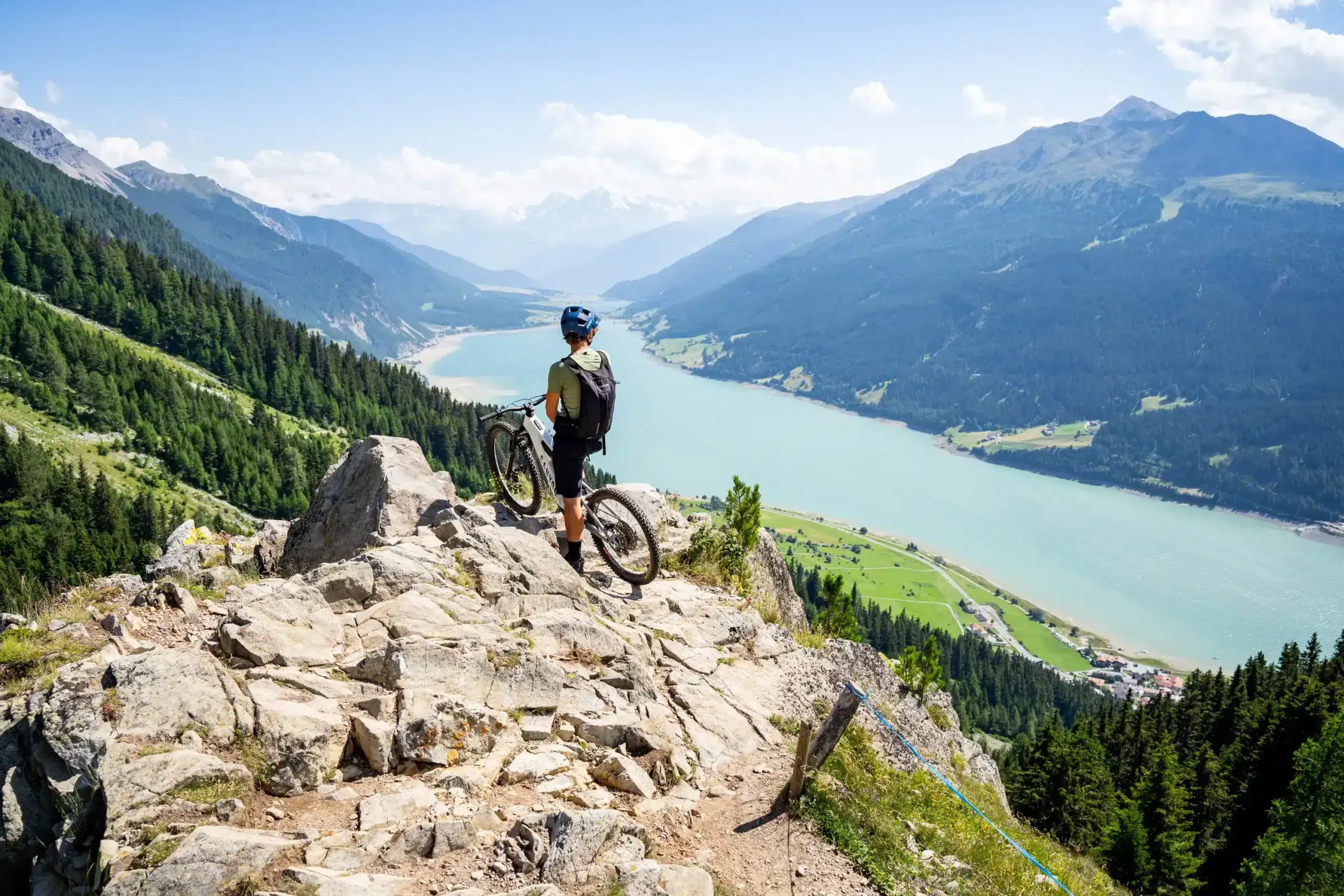 Eine Person mit einem Fahrrad steht auf einer felsigen Klippe mit Blick auf einen See und Berge in der Ferne.