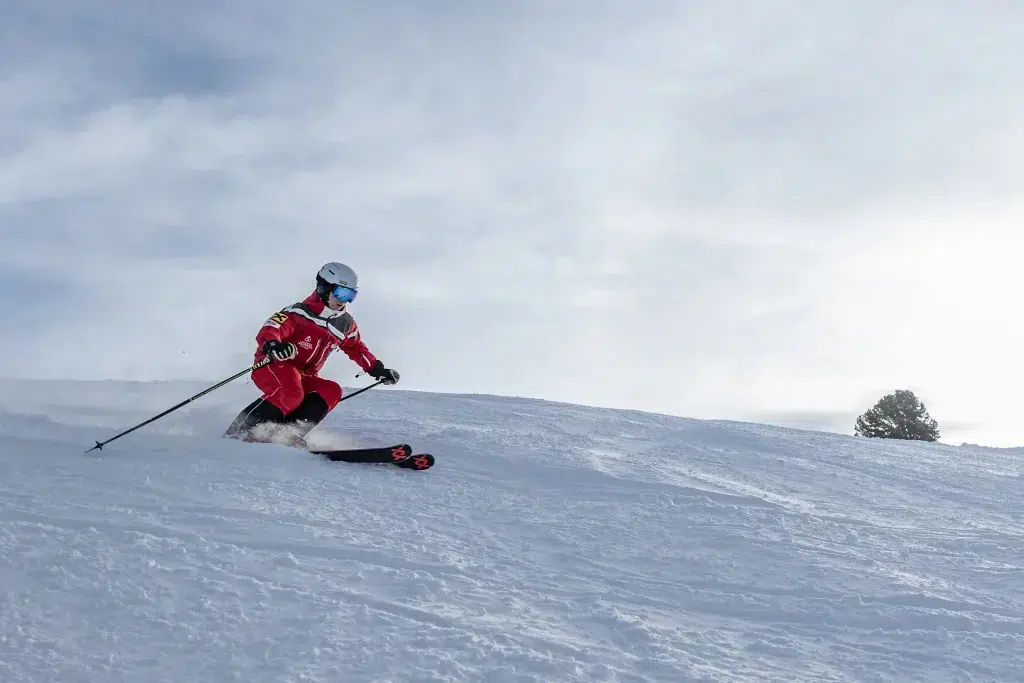 Erwachsene Skifahrerin in rotem Outfit fährt eine verschneite Piste unter wolkigem Himmel hinab.
