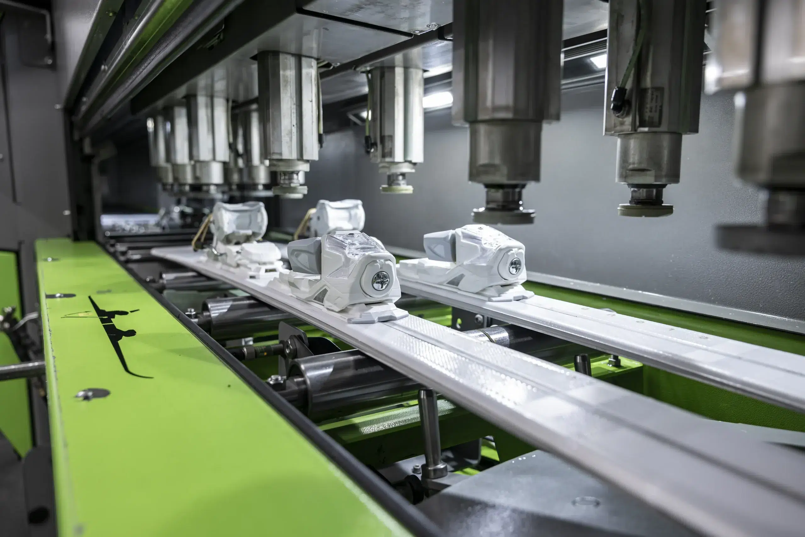 Weiße Skibindungen werden auf einer automatisierten Fertigungsstraße mit grünen Maschinenteilen montiert.
