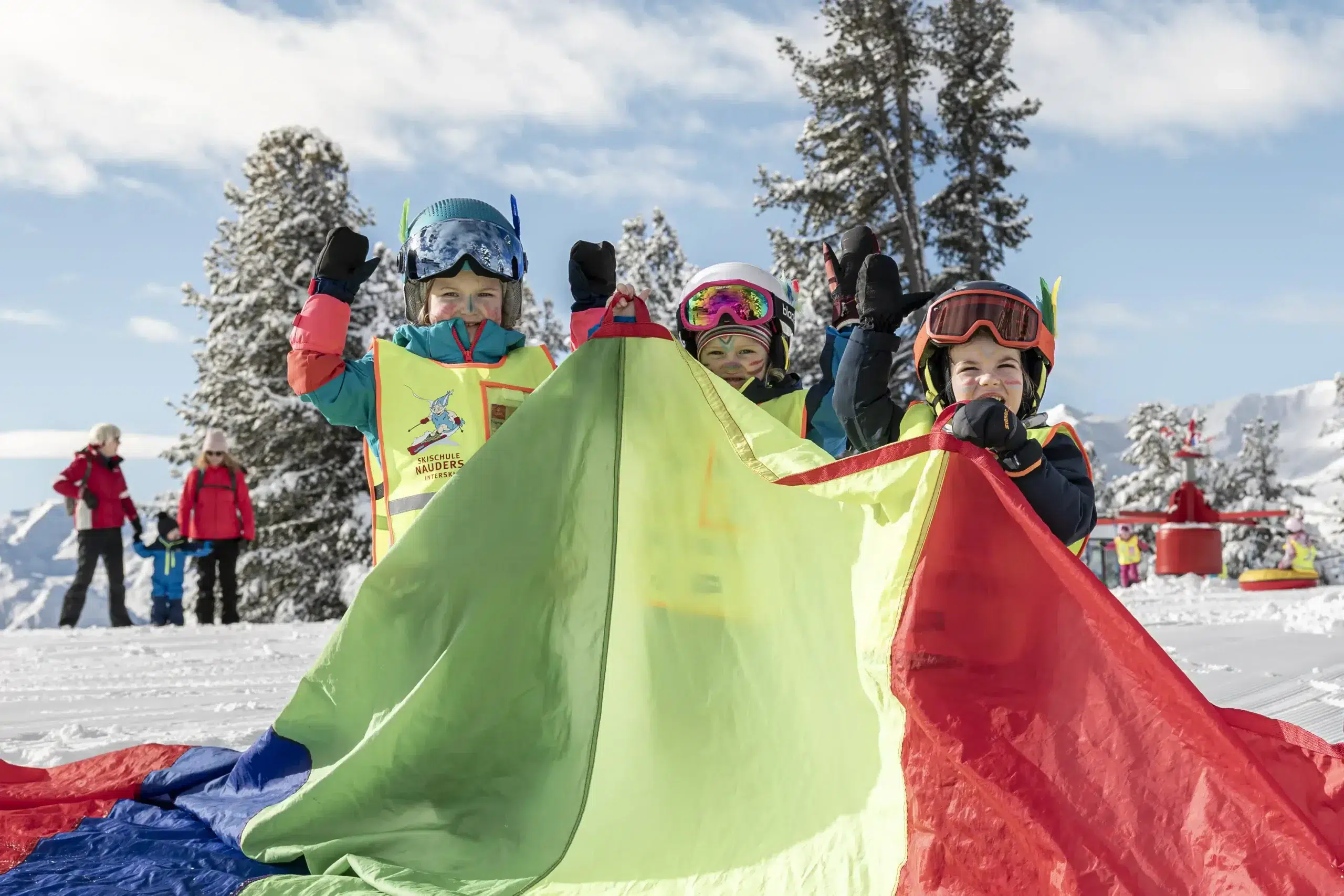 Drei Kinder in Skiausrüstung mit einem bunten Fallschirm auf einem verschneiten Berg.