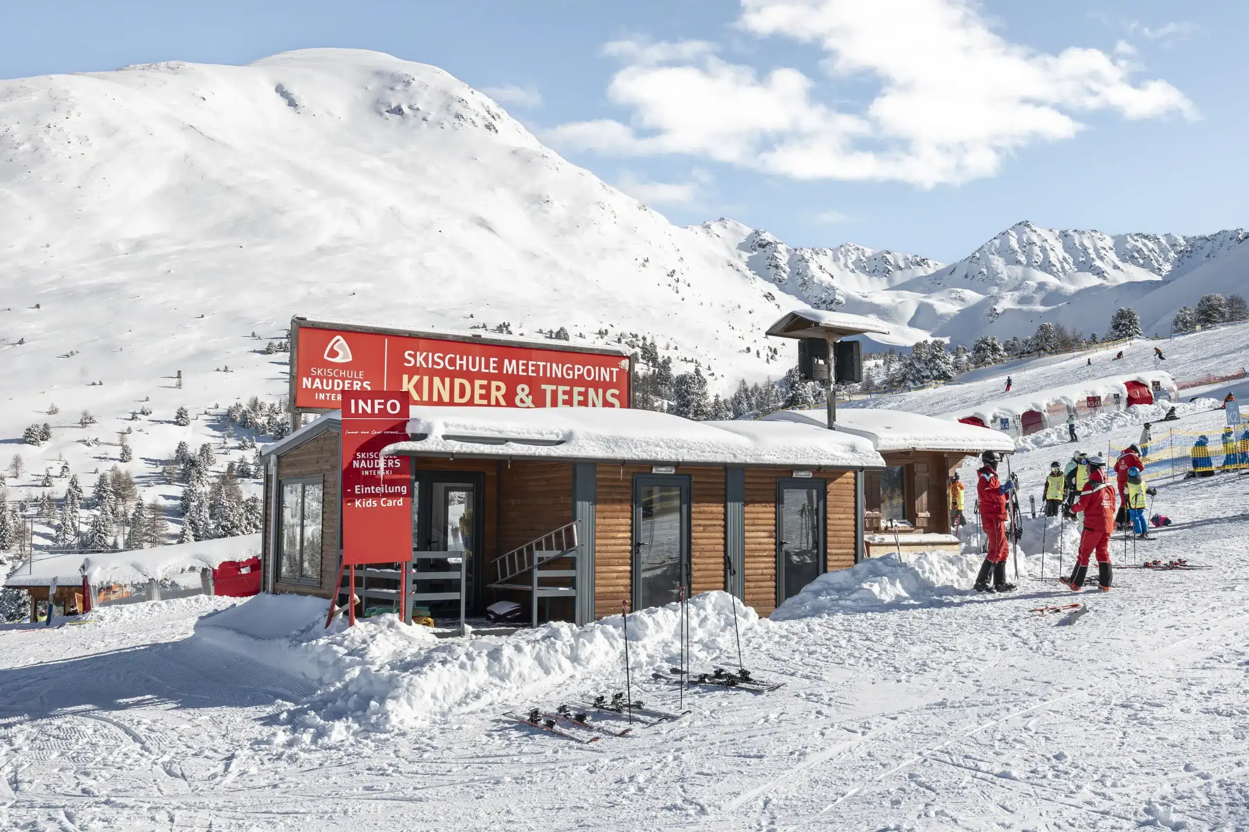 Skischultreffpunkt für Kinder und Jugendliche mit verschneiten Bergen und Skifahrern im Hintergrund.
