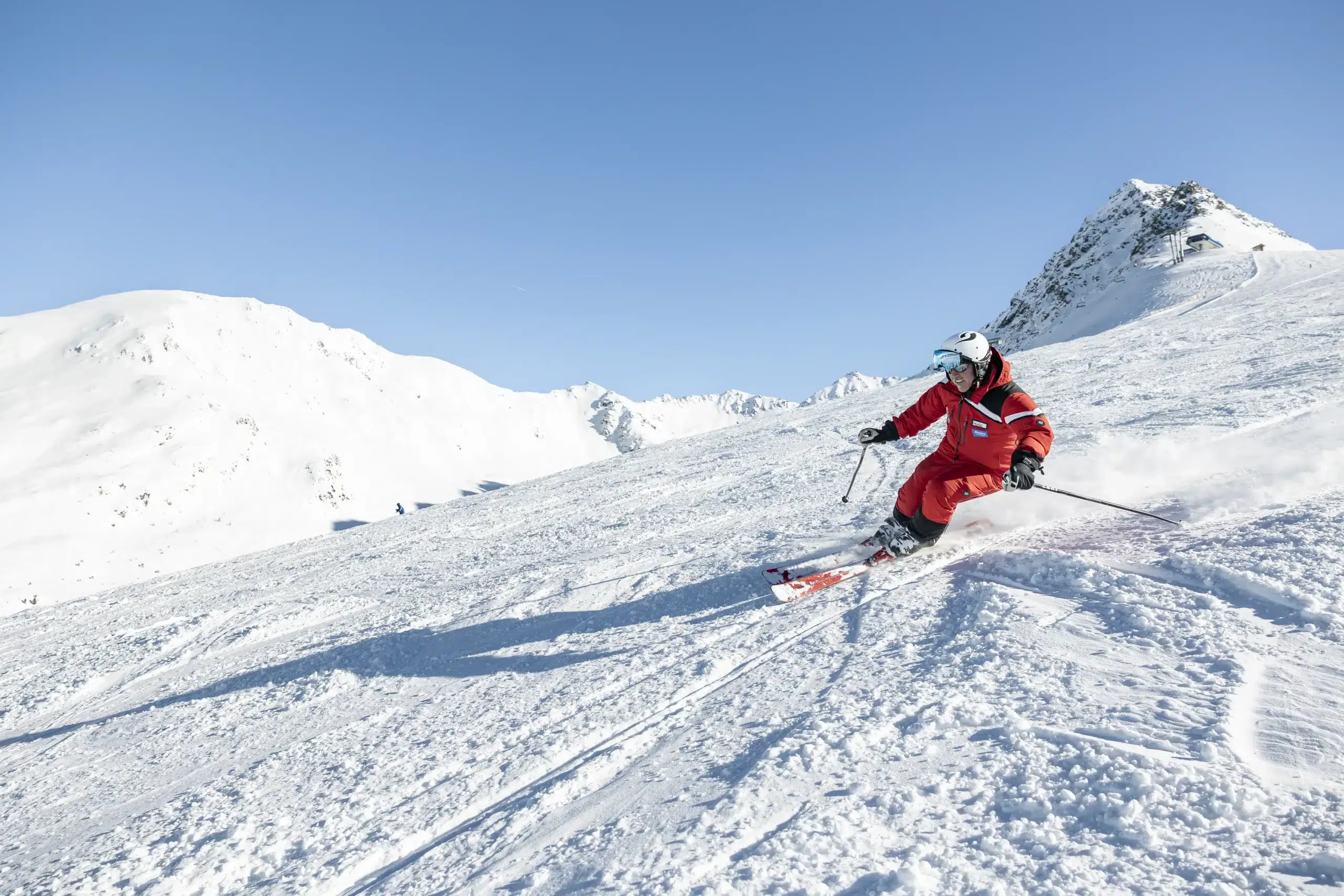 Erwachsene in rotem Anzug fährt die verschneite Piste bei klarem, blauem Himmel hinab.