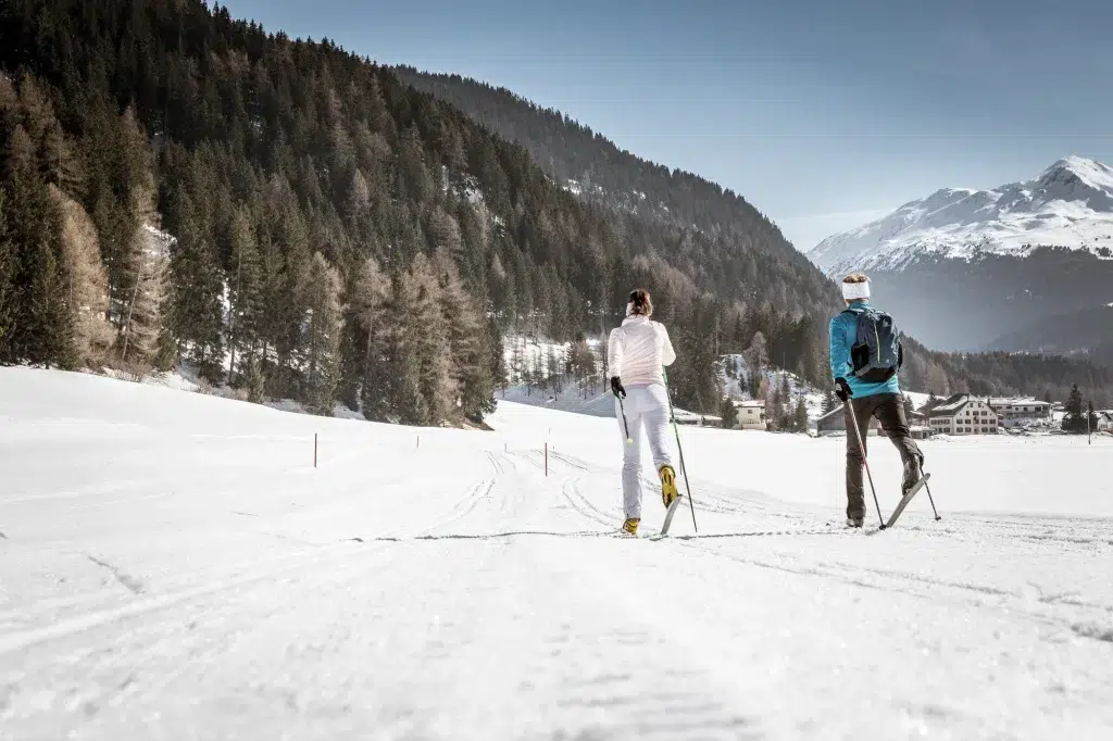 Zwei Personen beim Skilanglauf auf einer verschneiten Loipe mit Bergen und Bäumen im Hintergrund.